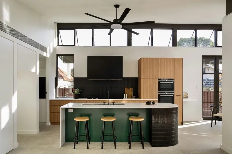 Open-plan kitchen with a black vertical-tile splashback, oak tall cabinets and a deep green island with a curved fluted end