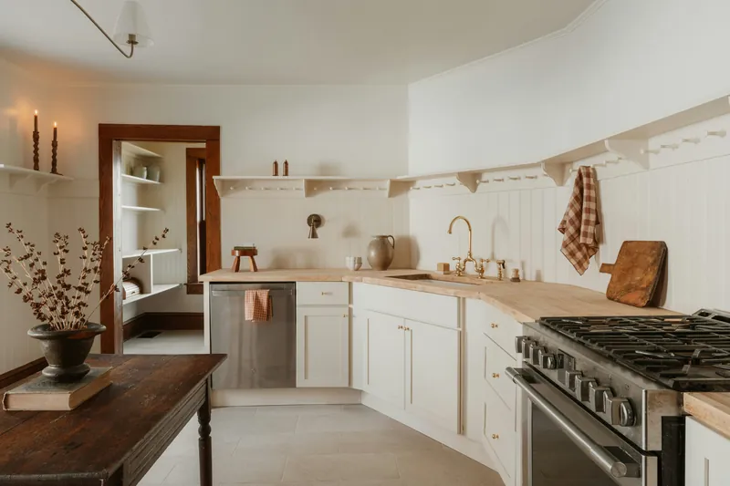 Cream beadboard kitchen with butcher-block counters, an unlacquered brass bridge tap and a peg-rail shelf along the wall