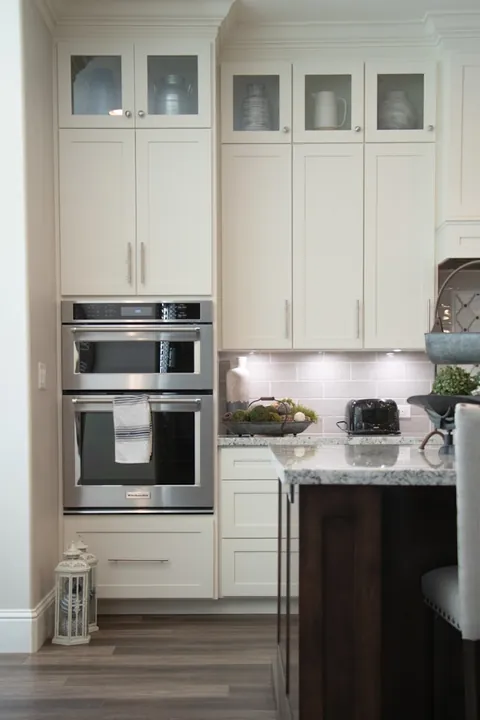 Cream cabinets with glazed upper doors flanking a stacked pair of stainless wall ovens above a drawer bank