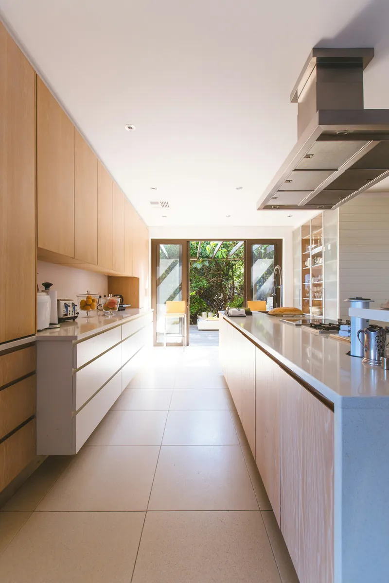 Long galley kitchen with oak tall cabinets down one side, a white run opposite and a stainless island hood overhead