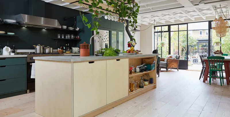 Deep green kitchen cabinets and open shelving beside a birch plywood island with a concrete worktop, opening to a garden