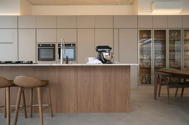 Floor-to-ceiling handleless cabinetry in warm taupe beside a walnut island with a marble top and lit glass display cabinets