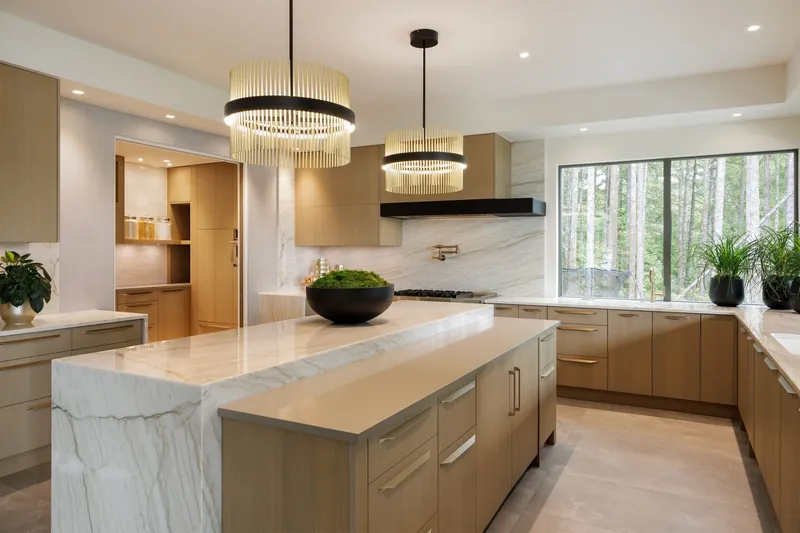Luxury kitchen in light oak with two islands, marble waterfall ends, brass drum chandeliers and a window onto forest