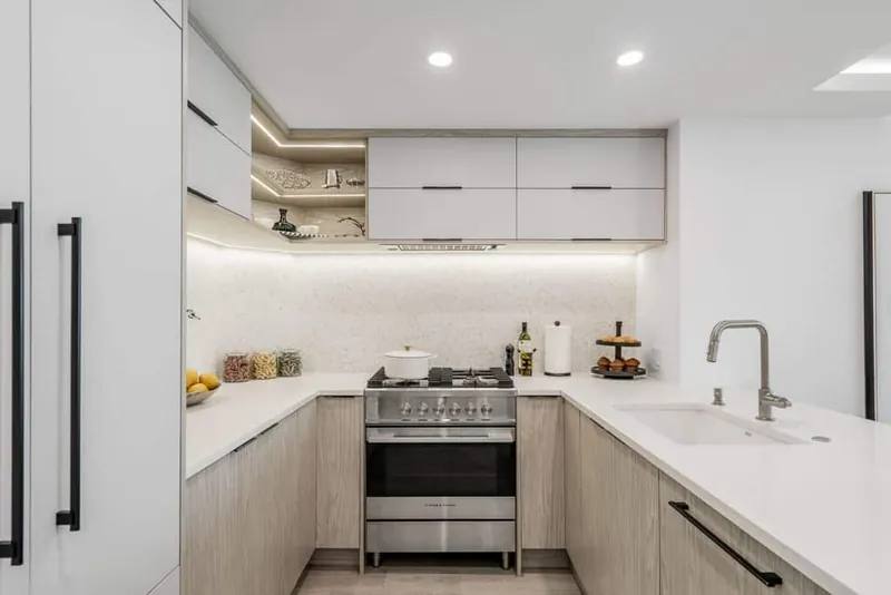 Small U-shaped kitchen with light oak lower cabinets, white handleless uppers with integrated lighting and white quartz counters