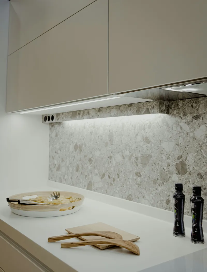 Warm LED strip lighting under greige wall cabinets washing down a speckled terrazzo-look stone splashback