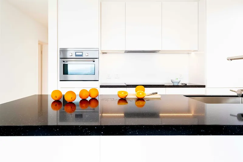 Minimal white handleless kitchen with speckled black quartz counters, an integrated cooktop and a stainless wall oven
