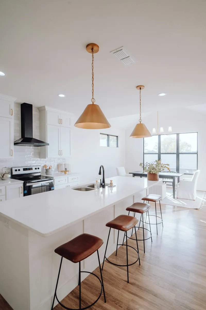 White shaker kitchen with a long quartz island, brass cone pendants, a black range hood and leather-topped bar stools